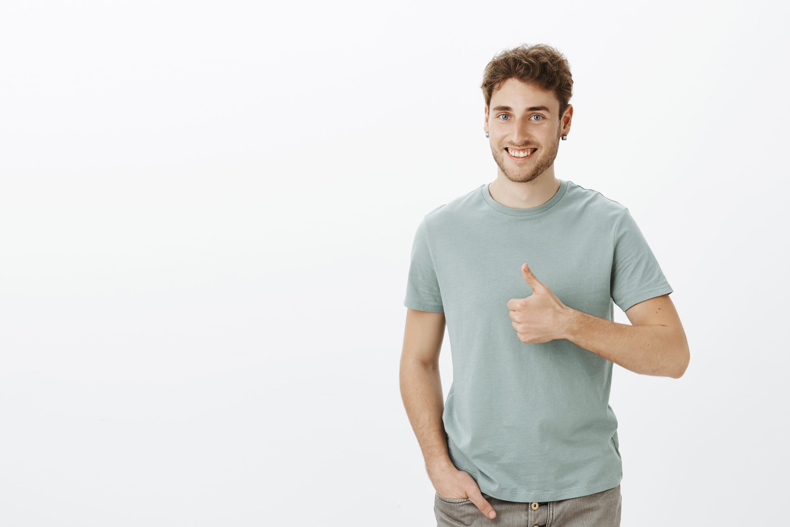 Positive happy young man in earrings, smiling broadly holding hand in pocket and showing thumbs up while being in good mood and liking new haircut of girlfriend, standing joyful over gray wall.